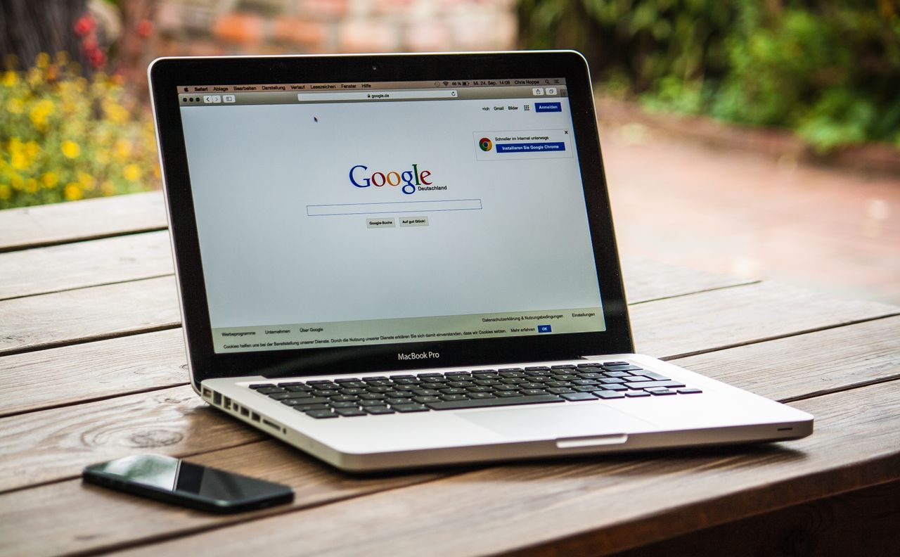 Un MacBook Pro montrant Google Search sur une table en bois en extérieur, à côté d'un smartphone.
